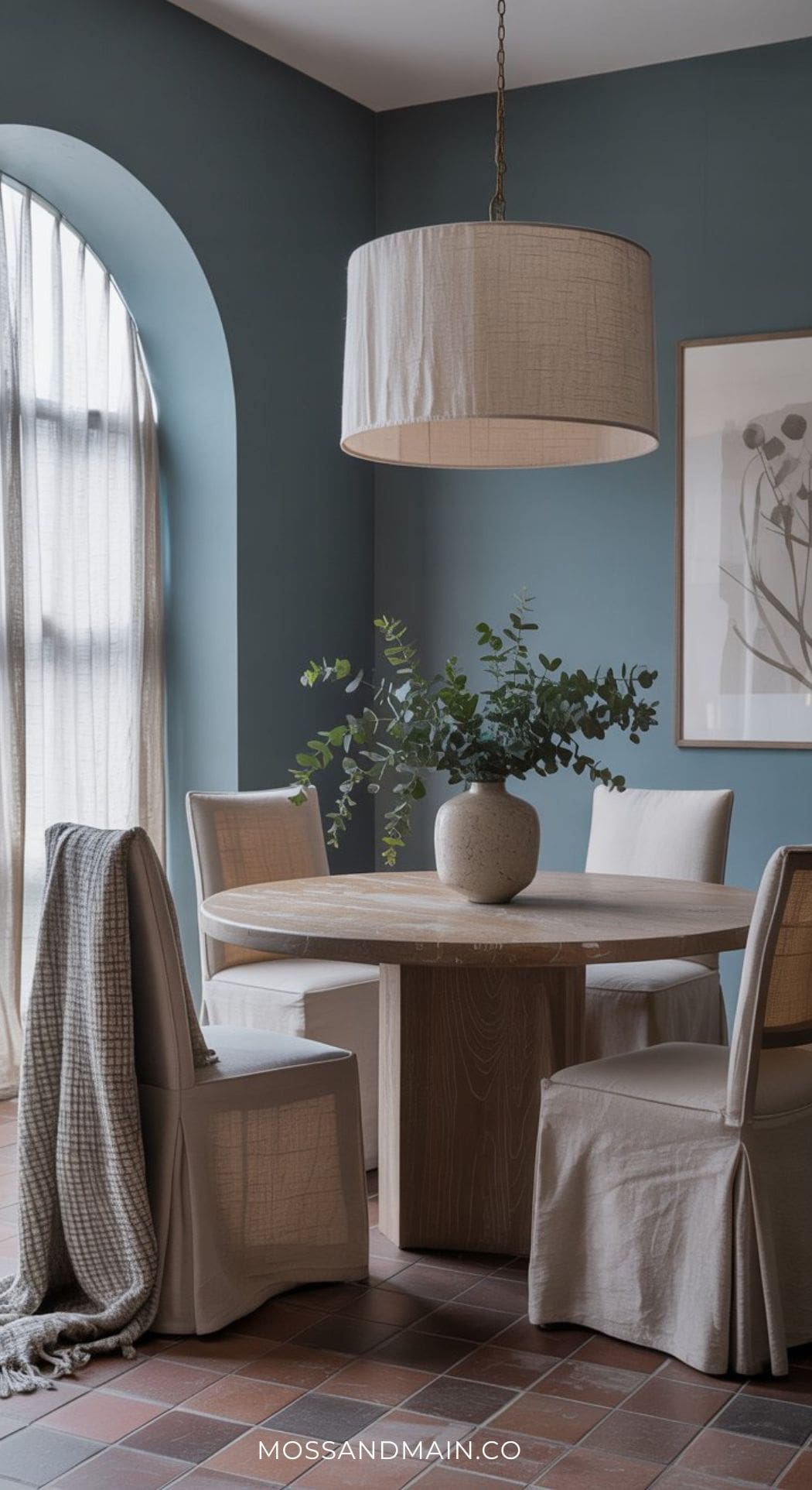 A cozy blue dining room with a round wooden table, four beige slipcovered chairs, a large pendant light, a vase with greenery, blue walls, an arched window with sheer curtains, and framed botanical art—perfect for inspiring Blue Dining Room Ideas.