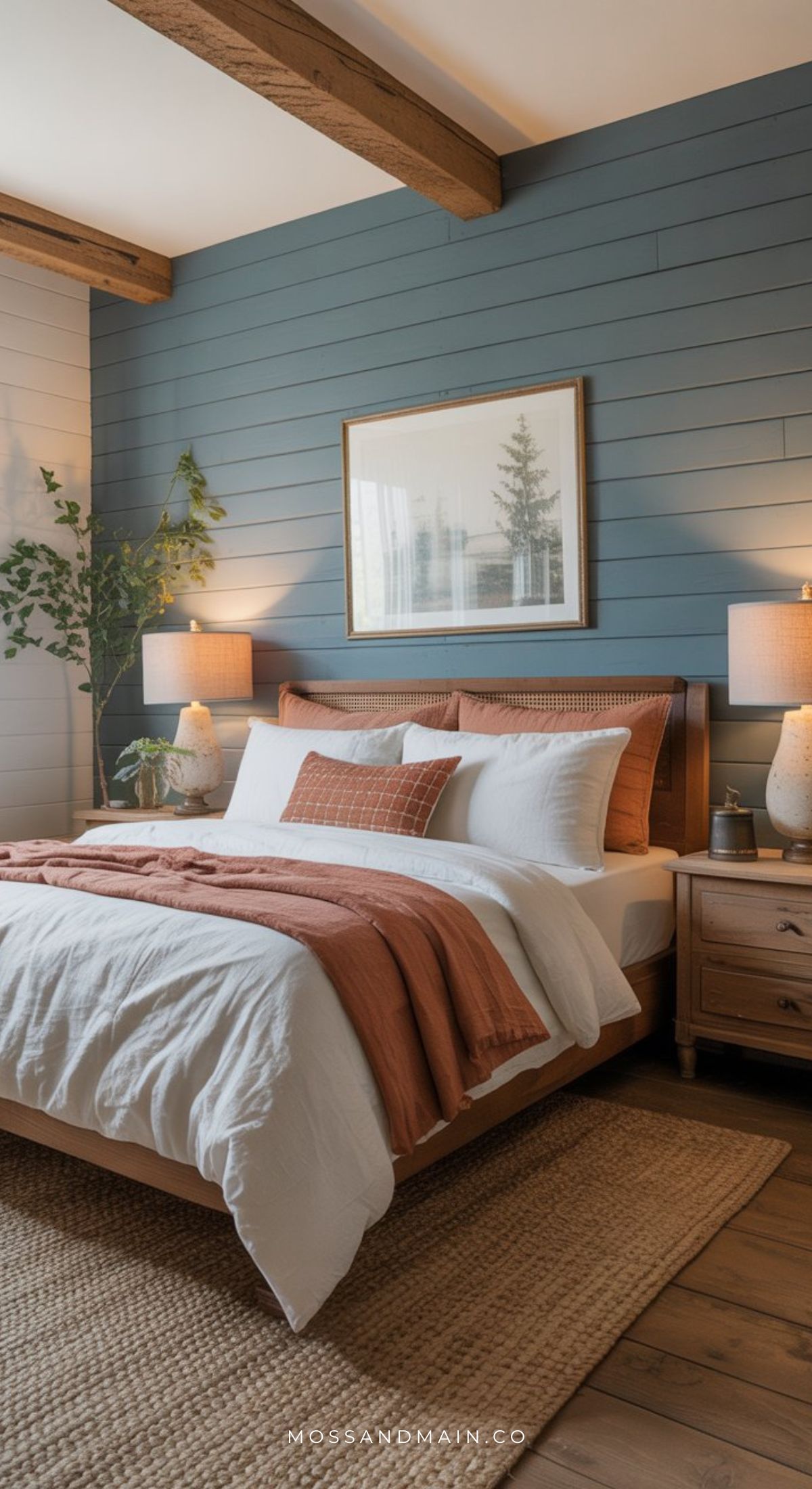 A cozy, earthy vibe fills this dusty blue bedroom, featuring a wooden bed, white and rust bedding, bedside tables with lamps, a woven rug, shiplap accent wall, framed art, and potted plants for decoration.
