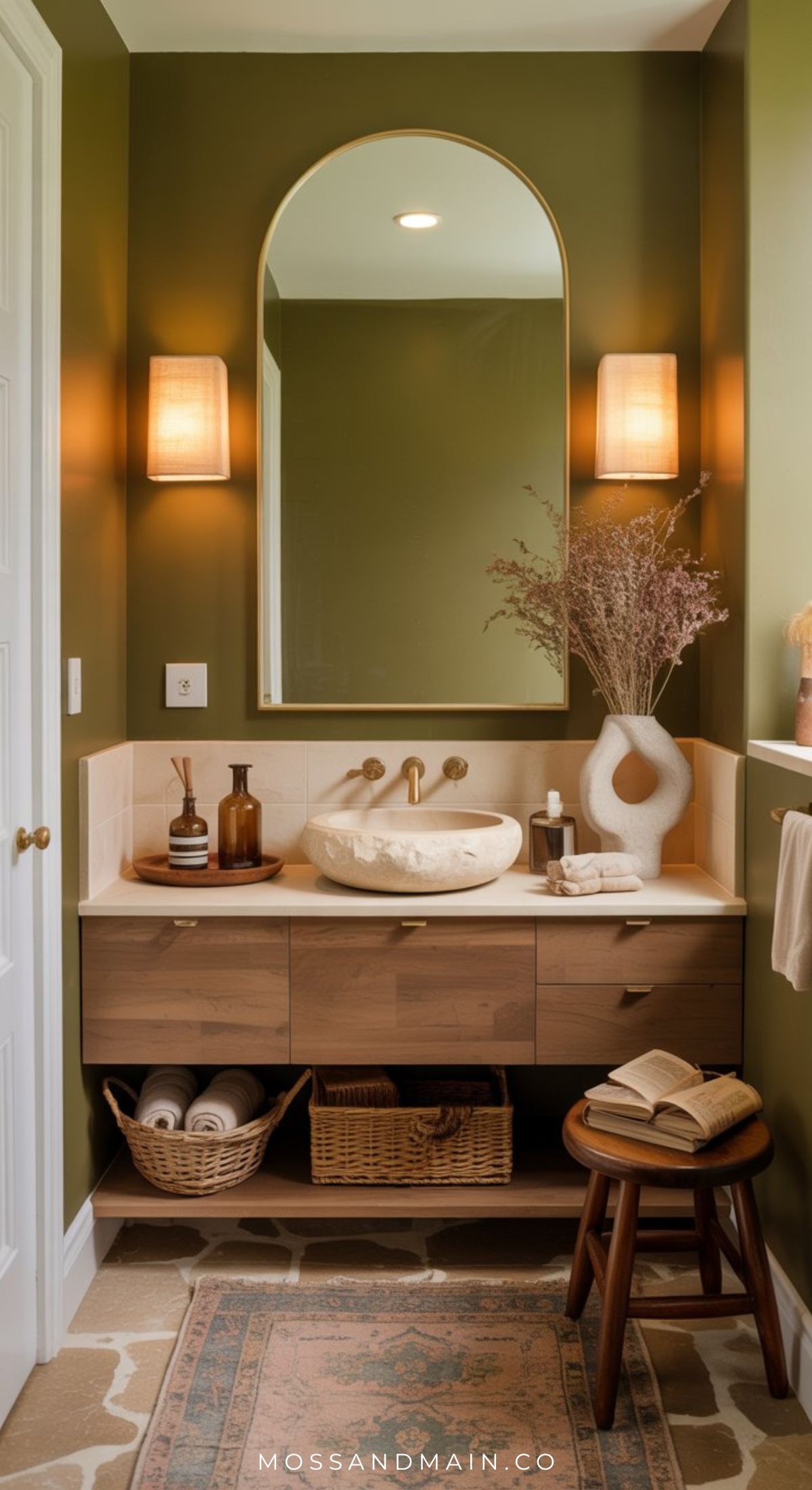 A small bathroom with olive green walls, a round-edged mirror, two wall sconces, a white stone sink on a wooden vanity, dried flowers, woven baskets below, and an open book on a stool.