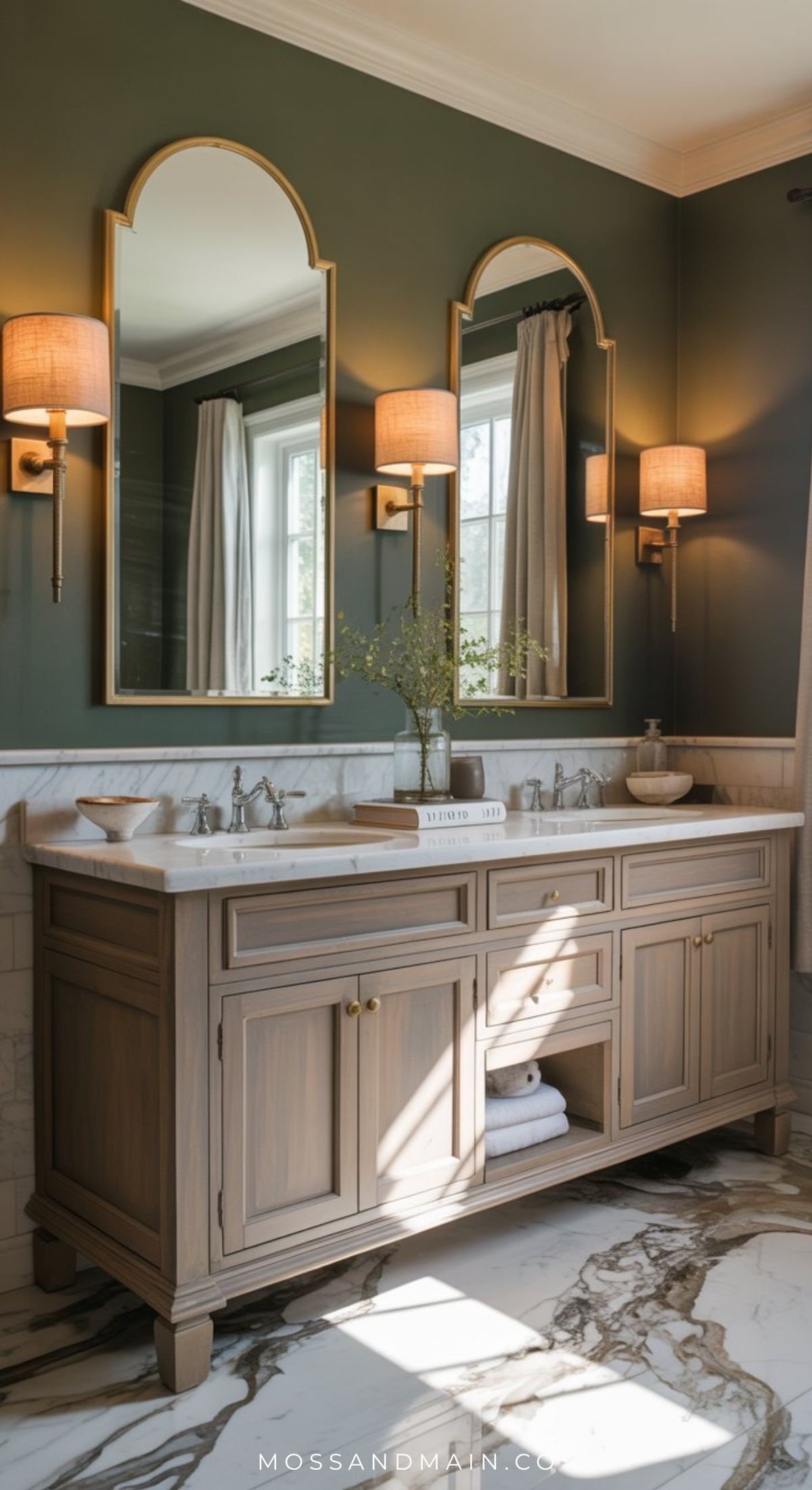 Elegant olive green bathroom with double vanity wood cabinets, marble countertop, two arched mirrors, wall sconces with beige shades, and a large window filling the space with light. Marble flooring completes this inspiring bathroom design.