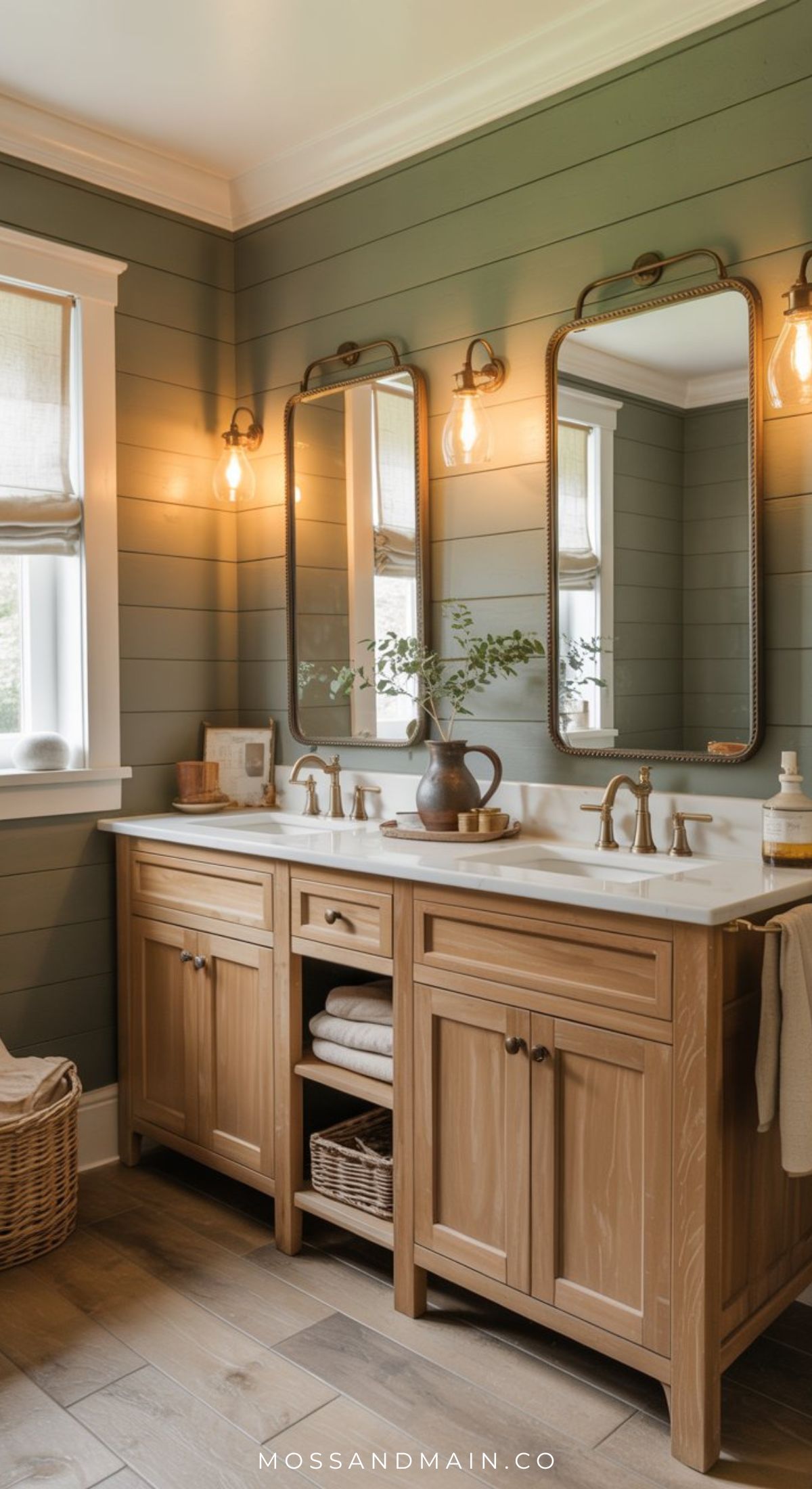A cozy bathroom with green shiplap walls, double wooden vanity, two mirrors, brass fixtures, woven basket, and soft ambient lighting from wall sconces. Towels and a vase with greenery sit on the countertop.