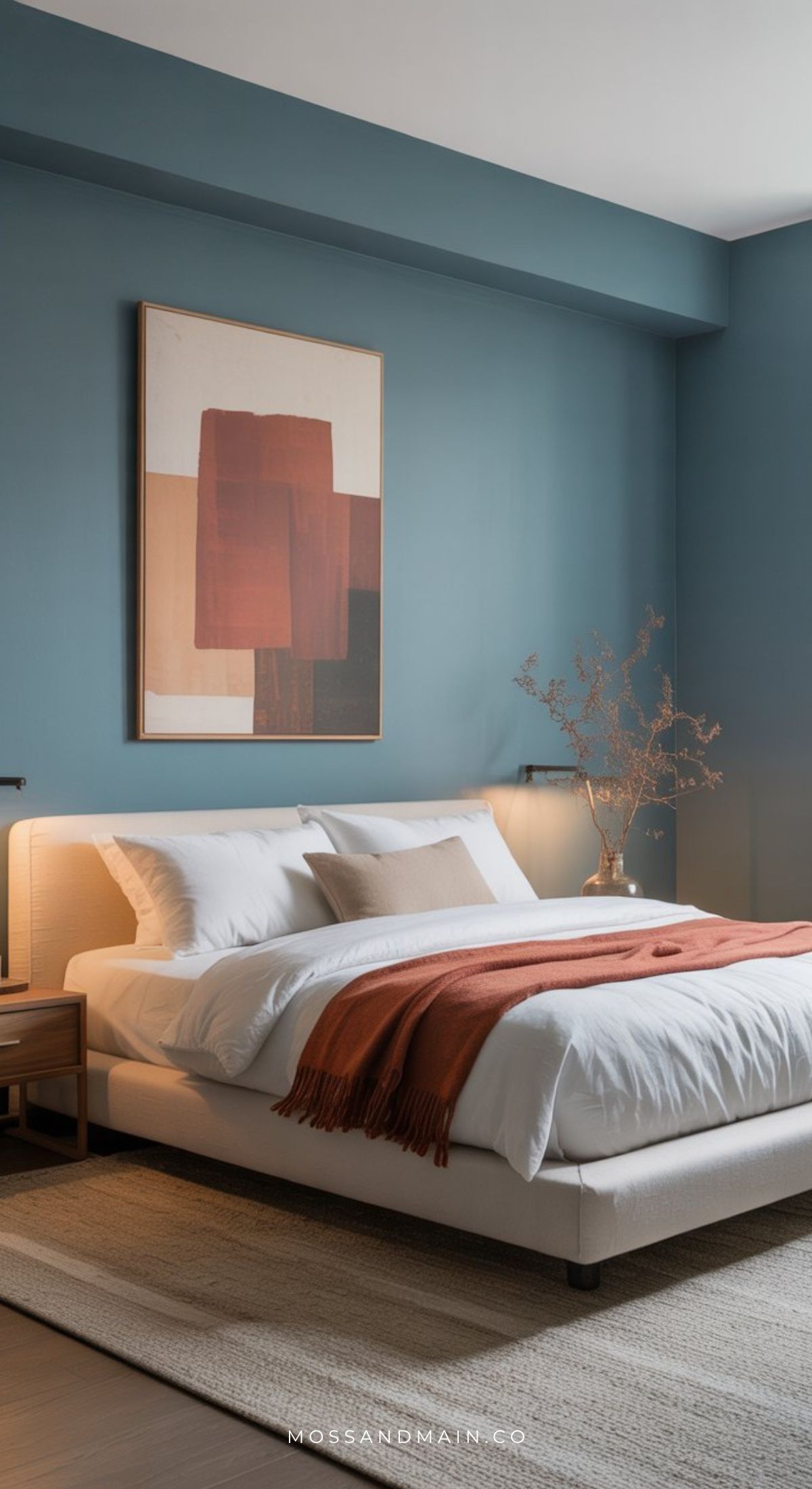 Modern bedroom with teal blue walls and a large abstract art piece above the bed, featuring cozy earthy vibes. White bedding and a rust-colored throw blanket add warmth, while natural light completes this Dusty Blue Bedroom look.