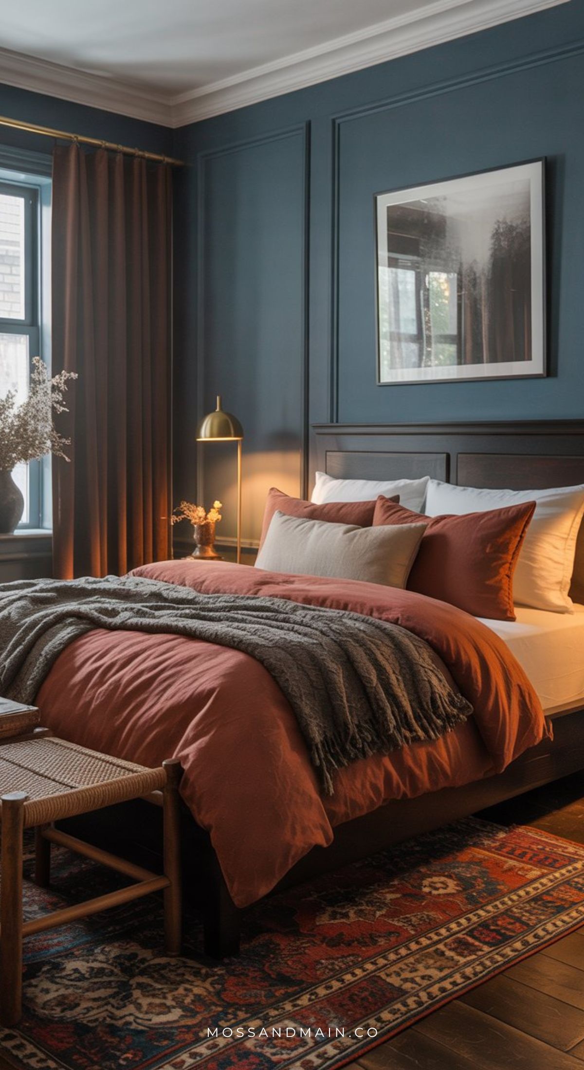 A cozy bedroom with dark blue walls, a wooden bed with rust-colored bedding and gray blanket, a vintage rug, a floor lamp, and earthy accents captures beautiful Rust Bedroom Ideas for those seeking a cozy earthy vibe.