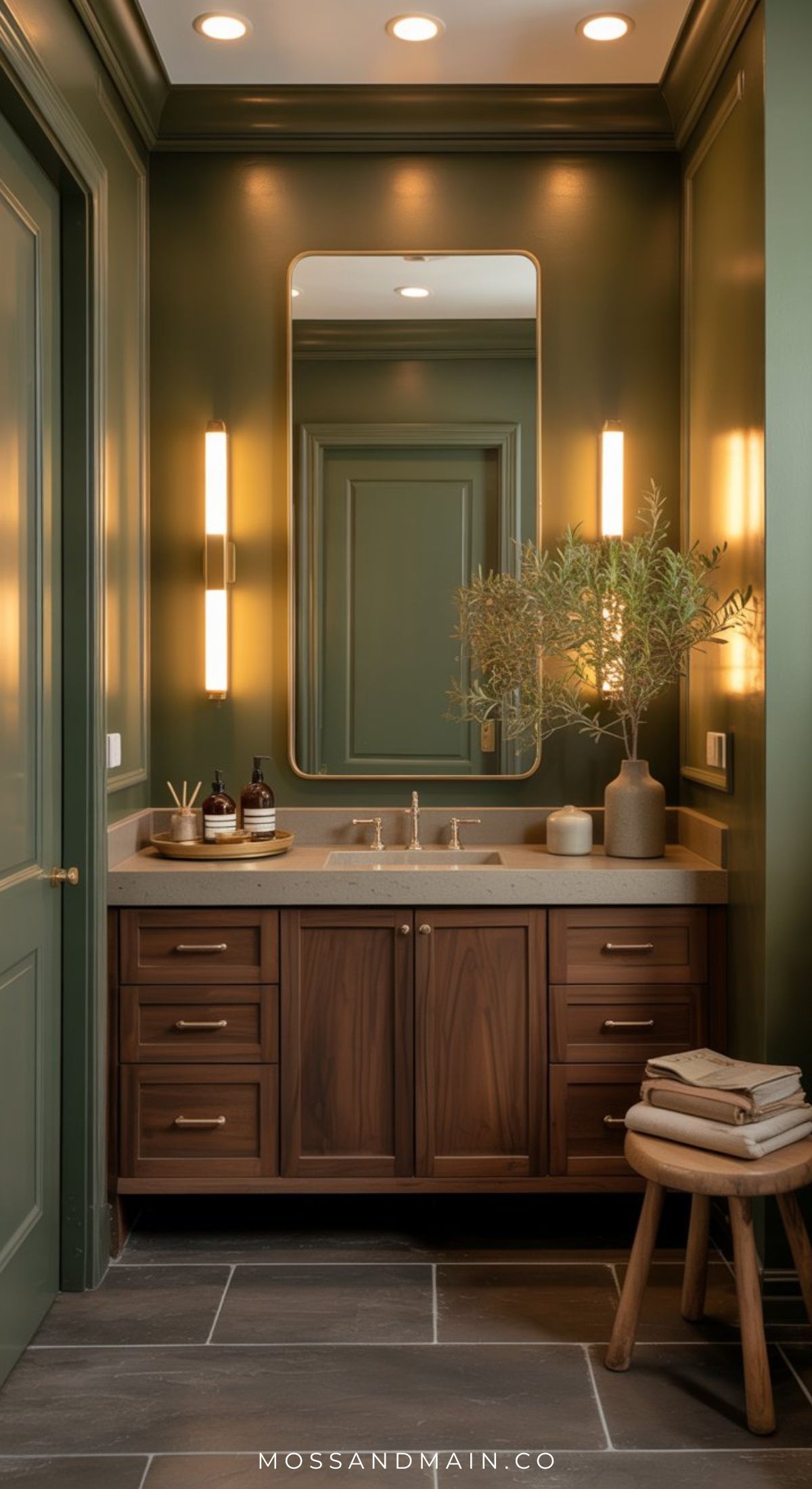 A stylish olive green bathroom with a wood vanity, rectangular mirror, modern sconces, potted plant, and stacked towels on a wooden stool—perfect inspiration for chic bathroom designs. Countertop holds soap dispensers and decor items.