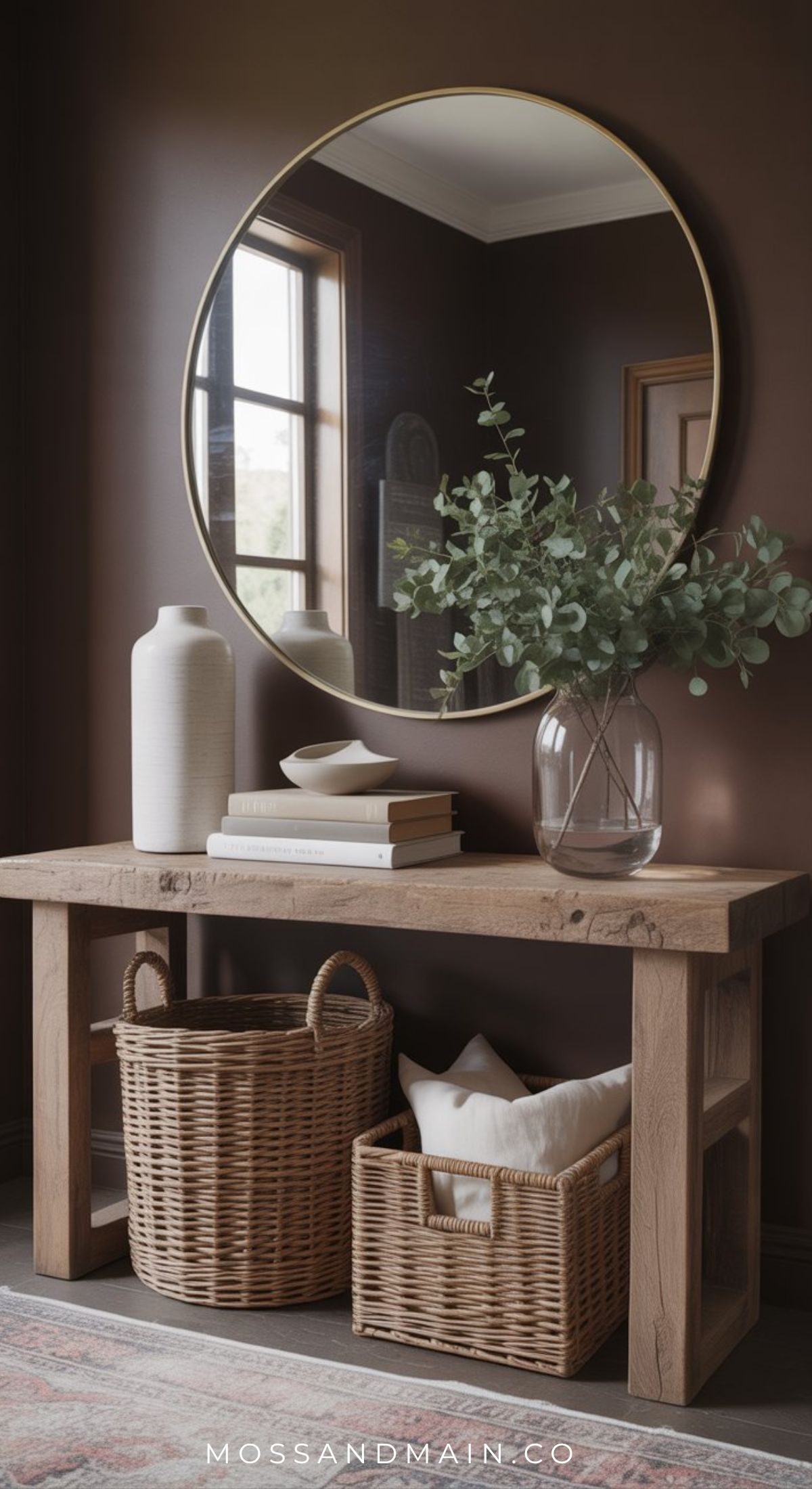 A wooden console table with two wicker baskets beneath, decorative books, a vase, and a glass jar with greenery on top offers effortless entryway charm. A large round mirror above reflects the window and adds to this warm entryway feel.