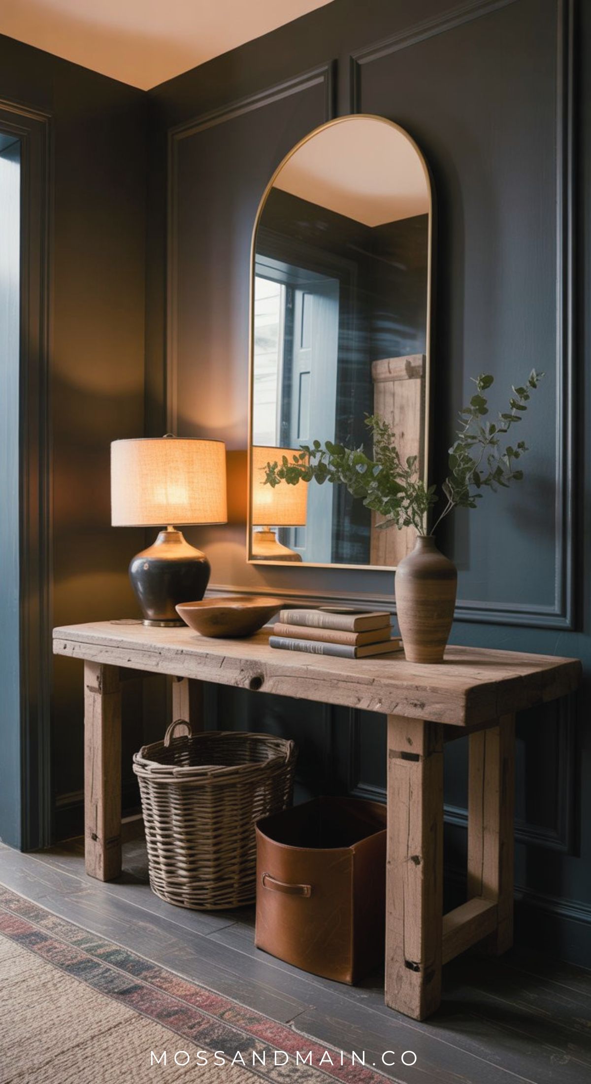 A rustic wooden console table creates a warm entryway, styled with books, a lamp, greenery in a vase, and a bowl. Set against a dark paneled wall and large arched mirror, it’s finished with a wicker basket and leather bin beneath for collected entryway charm.
