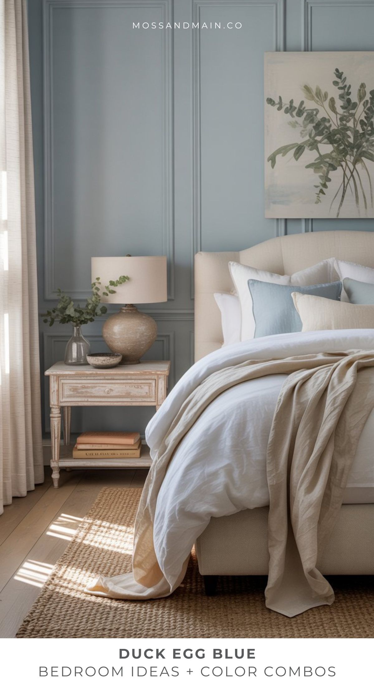 An elegant bedroom features a bed with white and beige linens, duck egg blue accent pillows, a matching upholstered headboard, a vintage nightstand with a vase and lamp, and a textured blue accent wall adorned with framed art and greenery.