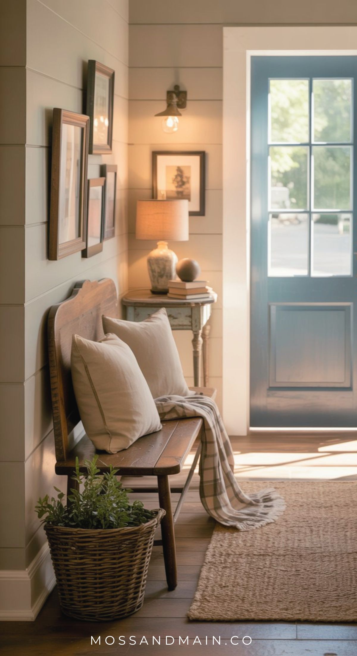 A cozy, warm entryway with a wooden bench, white cushions, and a plaid throw. A basket with greenery sits below. Framed art, a lamp, and decor rest on a table against paneled walls as sunlight streams through a blue door—effortless entryway charm.