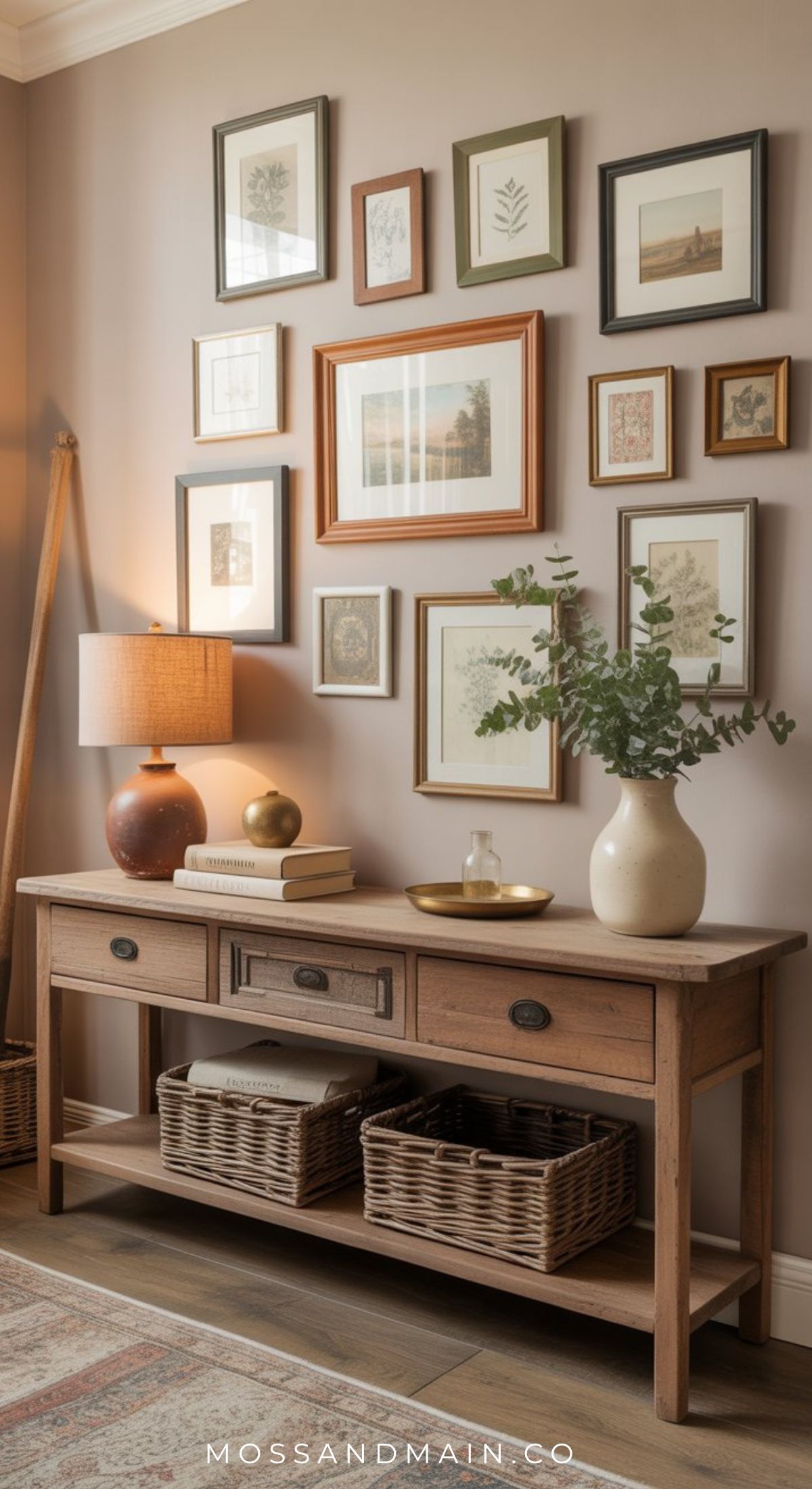 A wooden console table with baskets, books, and vases sits in a collected entryway against a wall of framed art. A glowing table lamp and a leafy branch in a vase add warmth to the cozy, stylish room.