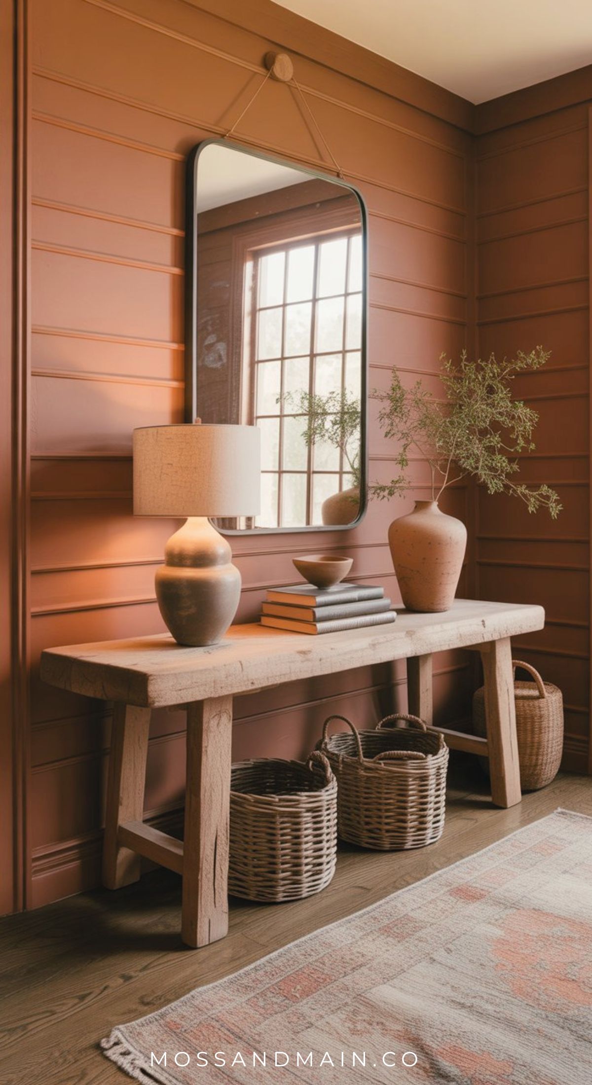 A wooden console table with a lamp, books, ceramics, and a plant creates an effortless entryway against a rust-colored paneled wall below a mirror. Woven baskets sit underneath as sunlight streams through the window reflected above.