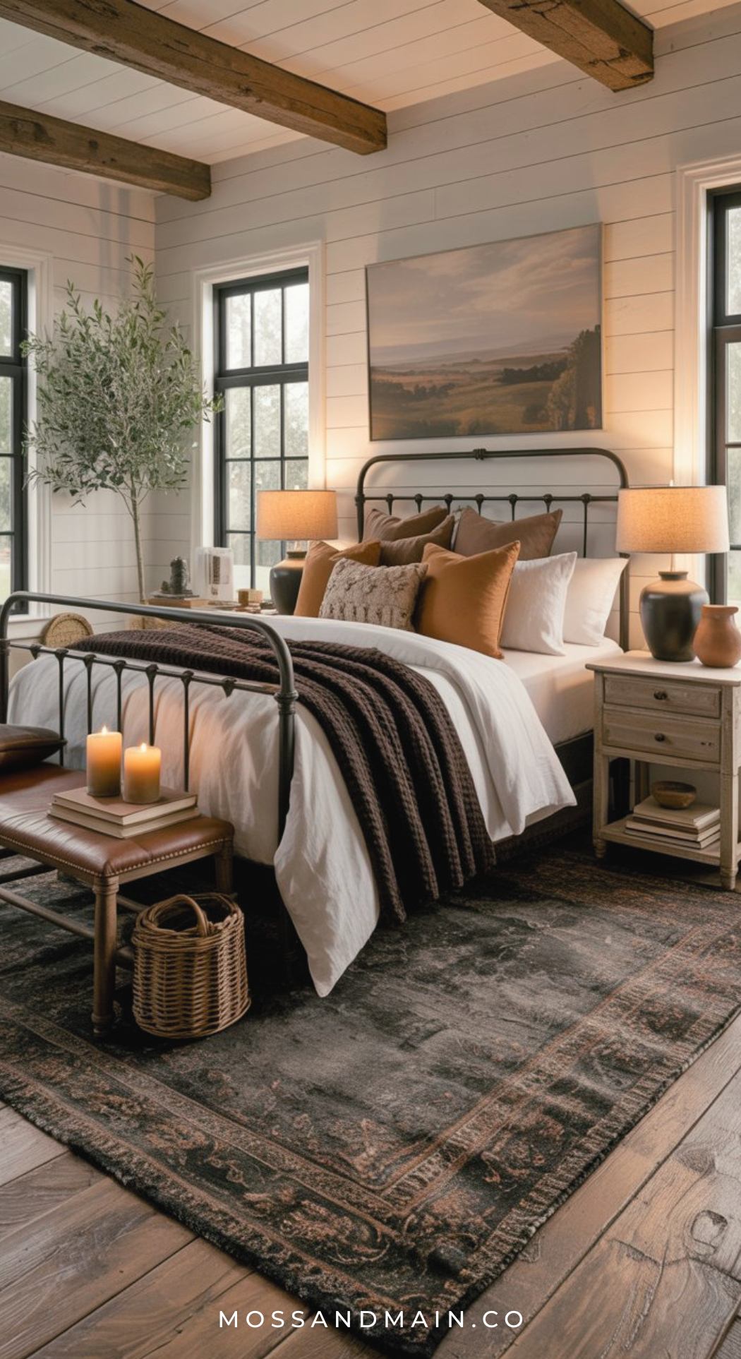 A cozy farmhouse bedroom with a metal bed frame, layered bedding in white and brown tones, rustic nightstands, warm lamps, an area rug, wooden beams, candles, and large windows offers cozy luxe bedrooms inspiration for brown bedroom ideas.