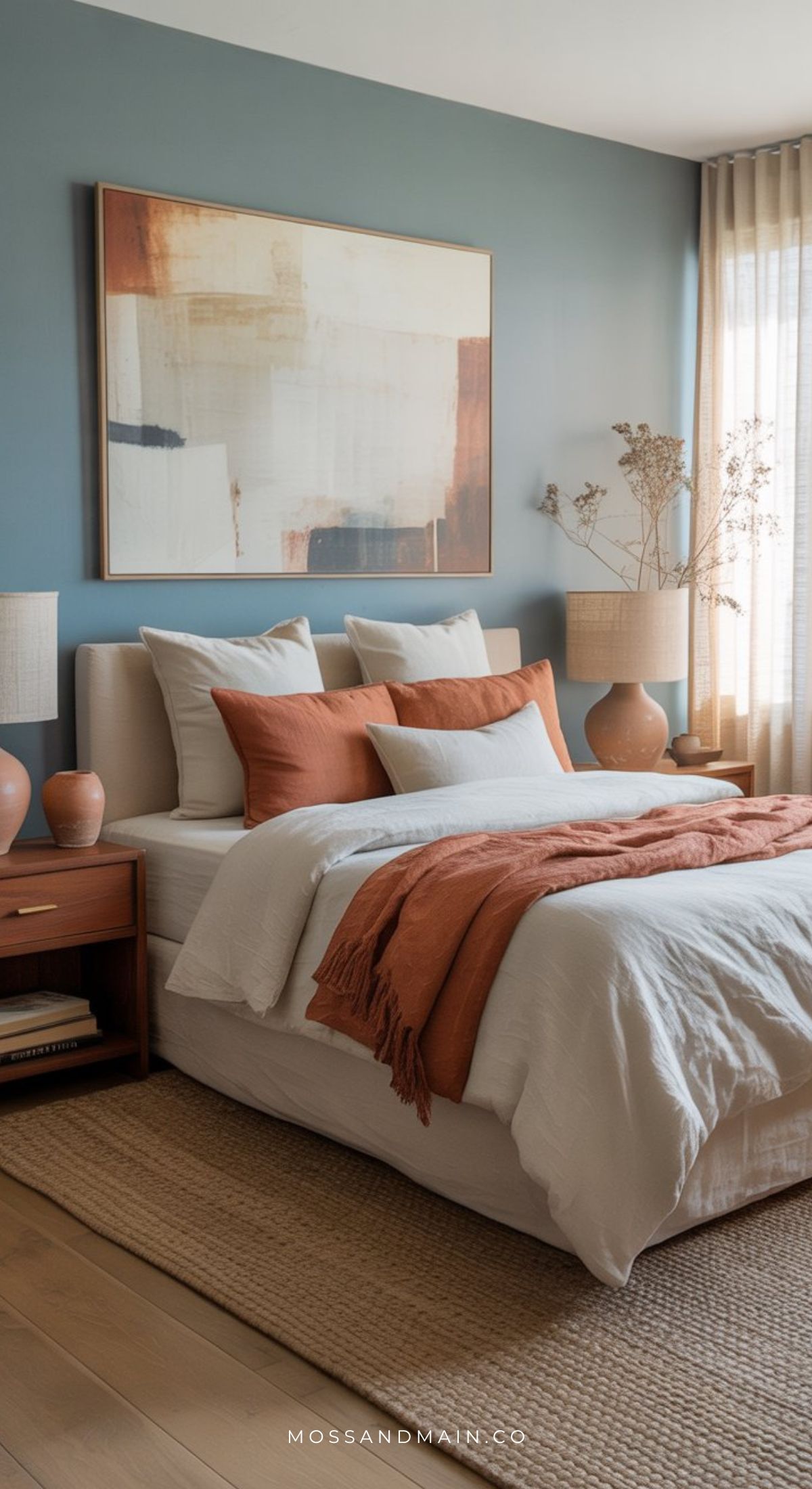 A modern dusty blue bedroom with a bold accent wall, abstract art, a neatly made bed with white and rust-colored bedding, two bedside tables with matching lamps, and natural light streaming through sheer curtains for an earthy vibe.