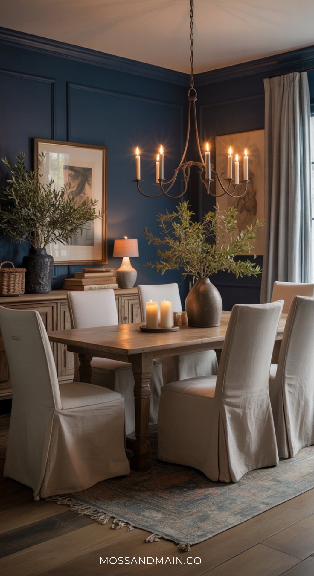 A cozy, moody dining room with navy blue walls, a wooden table surrounded by six white slipcovered chairs, candles, green branches in vases, warm chandelier lighting, framed art, and natural light from a window with beige curtains.