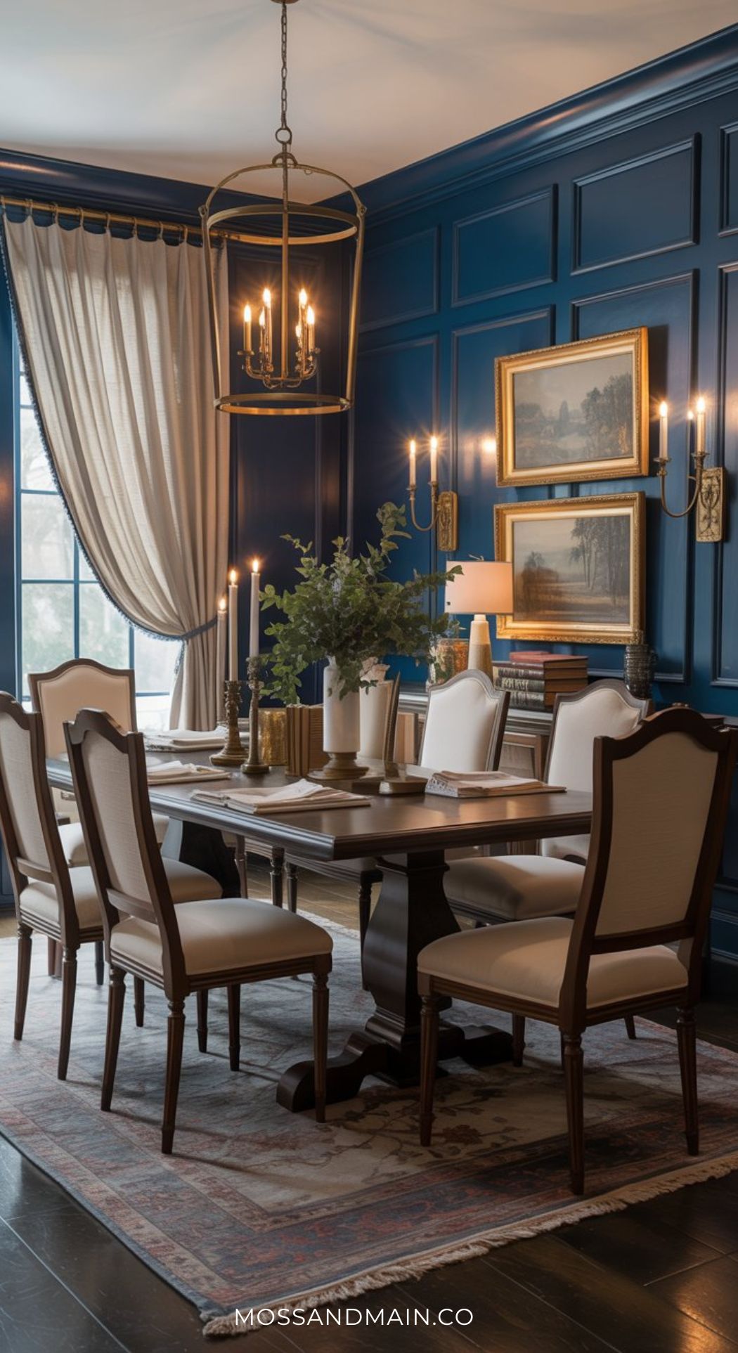Elegant, moody dining room with navy blue paneled walls, a wooden table surrounded by cream-upholstered chairs, gold-framed art, a classic chandelier, wall sconces, tall curtains, and a rug on dark wood floors.