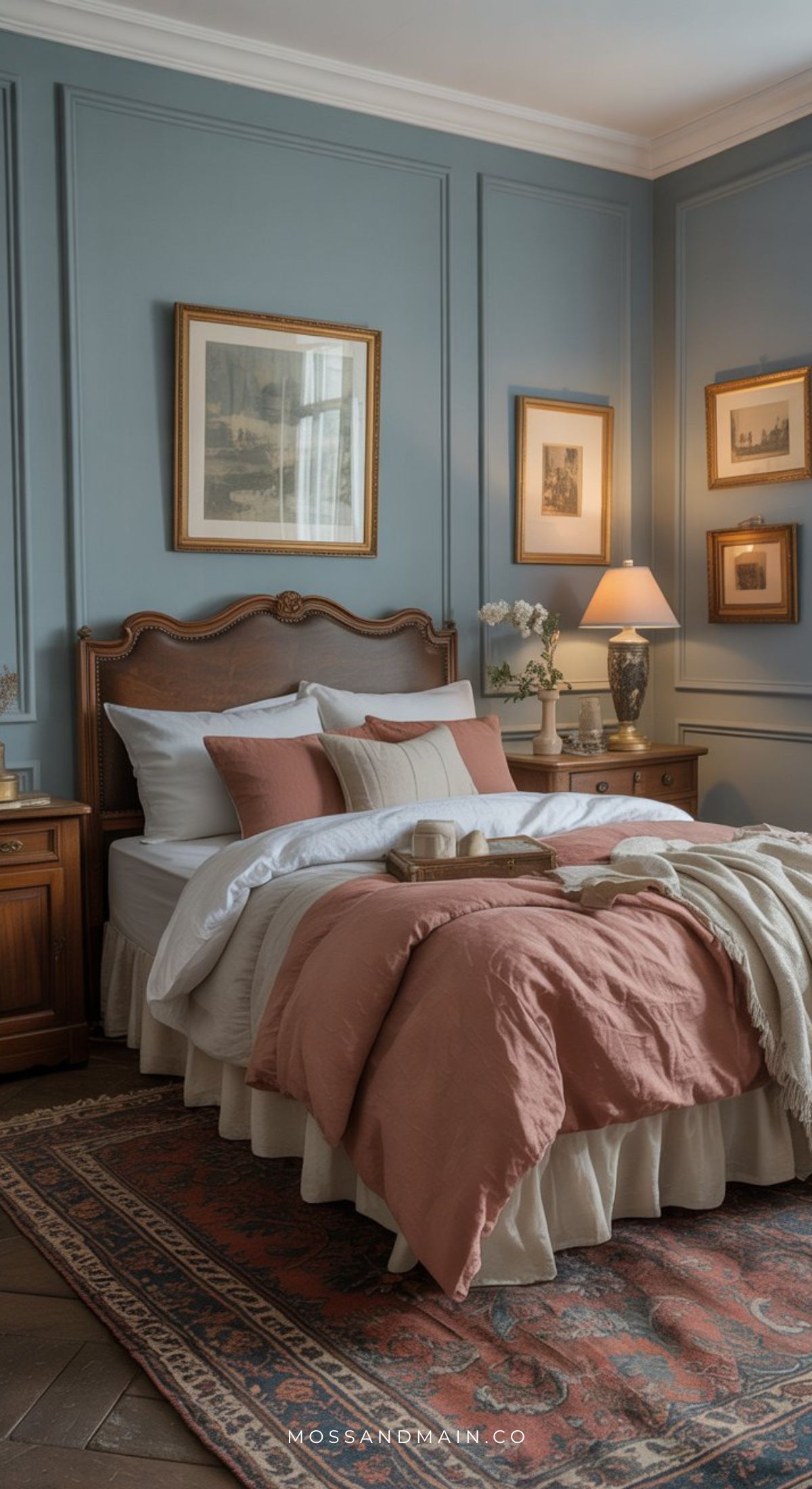 A cozy earthy bedroom with blue paneled walls, a wooden bed with white and blush bedding, matching pillows, two wooden nightstands, framed art, fresh flowers, a table lamp, and a patterned rug for the perfect dusty blue bedroom retreat.